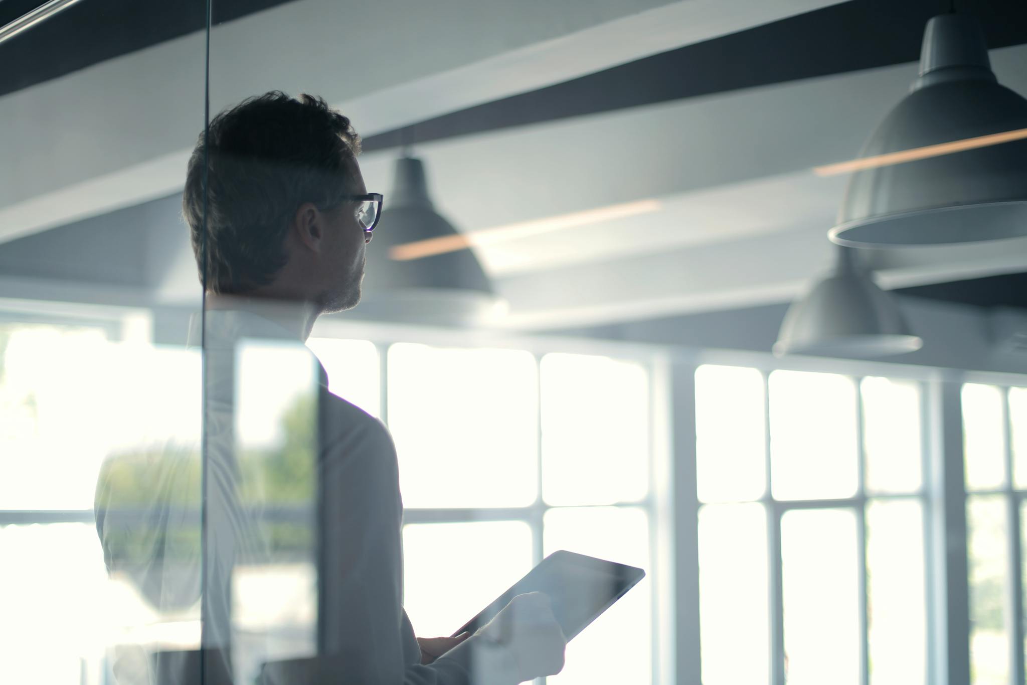 A businessman holding a tablet in an office, looking thoughtfully through a glass wall.